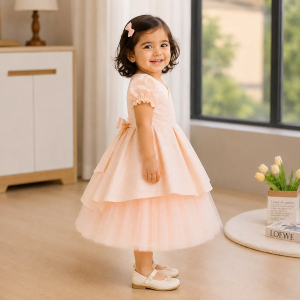 A young toddler girl wearing a light peach-pink tiered tulle dress with puff sleeves and cream-colored Mary Jane shoes, standing in a bright, modern room.