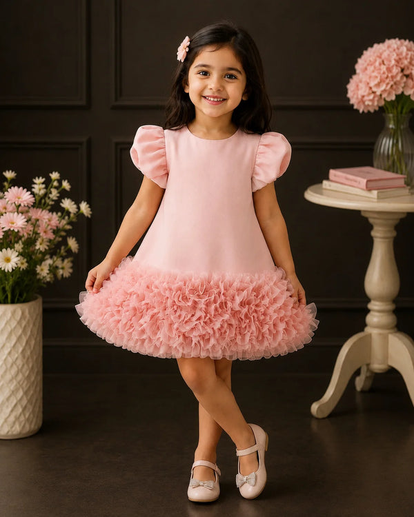A young girl with dark hair wearing a light pink dress with puffy short sleeves and a ruffled tulle skirt, standing in a decorated room with dark paneled walls, flowers, and a small table.