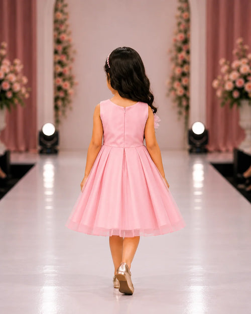 A young girl with long dark hair is seen from behind, walking down a runway wearing a sleeveless light pink dress with a pleated skirt and metallic gold shoes.