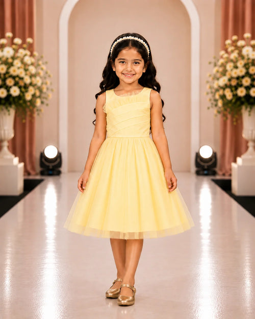 A young girl with long dark hair wearing a pale yellow sleeveless tulle dress and gold shoes, standing on a runway in a decorated indoor venue with floral arrangements and spotlights.