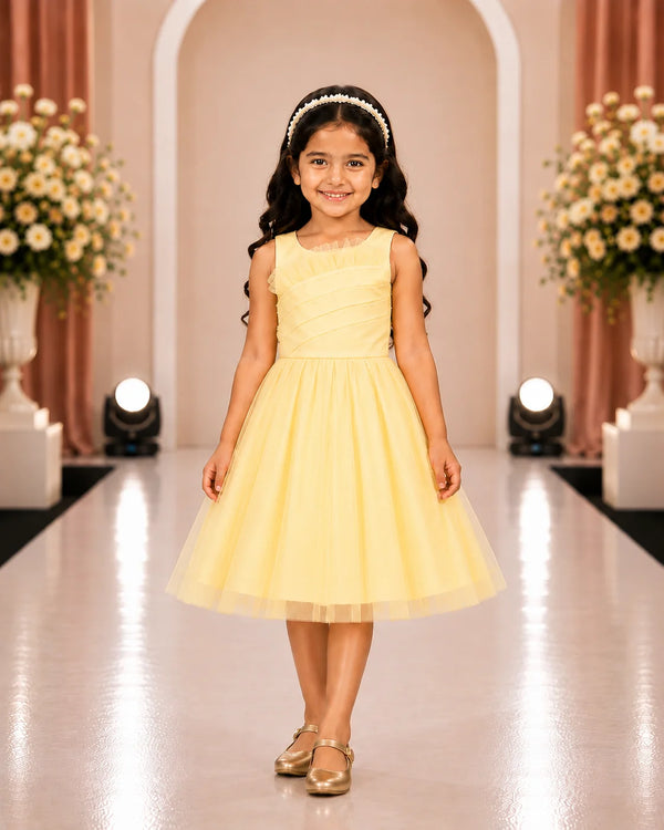 A young girl with long dark hair wearing a pale yellow sleeveless tulle dress and gold shoes, standing on a runway in a decorated indoor venue with floral arrangements and spotlights.