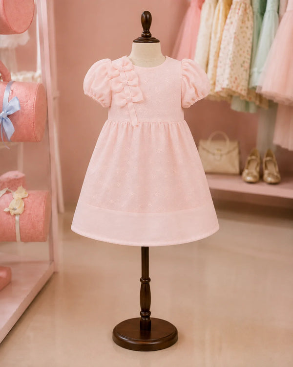 A light pink girls' dress displayed on a dress form mannequin in a boutique setting. The dress features puff sleeves, a round neckline, and a bow detail on the chest.