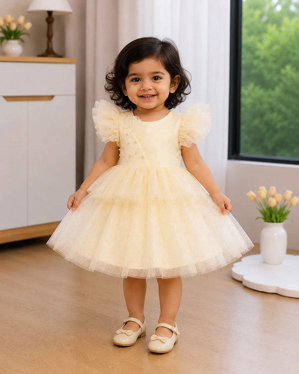 A toddler girl wearing a pale yellow tulle dress with puff sleeves and cream ballet flats, standing in a bright, modern room with wooden floors and a window.