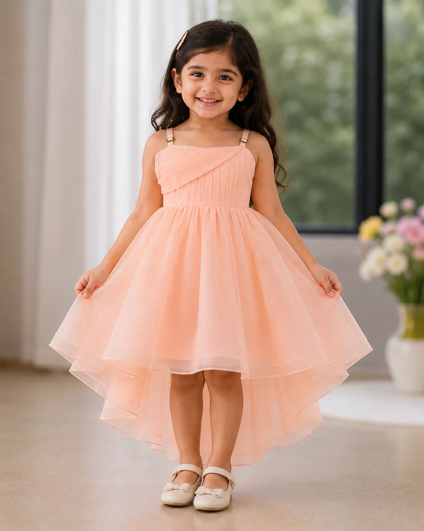 A young girl with long dark hair stands indoors wearing a sleeveless peach-pink high-low party dress with thin shoulder straps and a flared tulle skirt. She is posed in front of a softly blurred window and flowers.