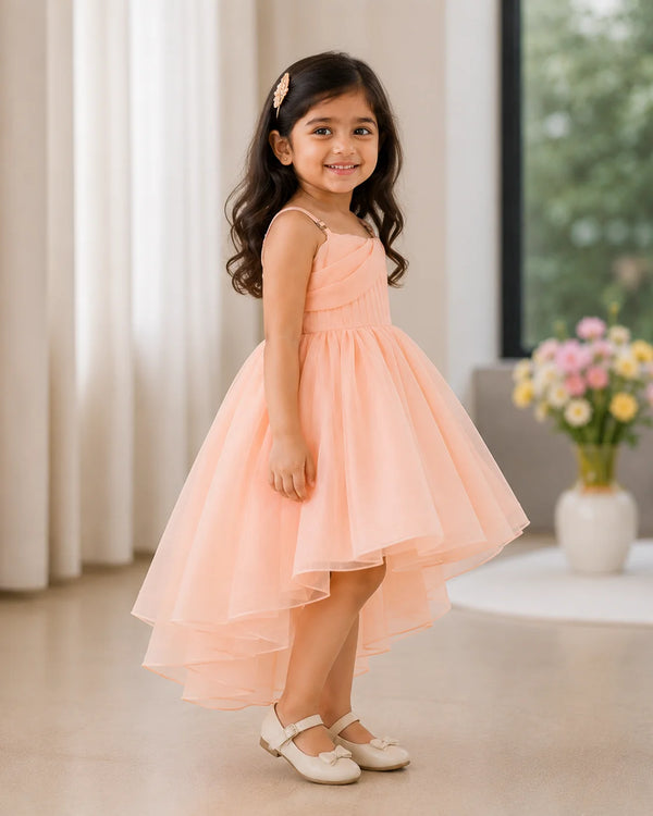 A young girl with long dark hair stands indoors wearing a peach high-low party dress with thin straps and a full tulle skirt, paired with white Mary Jane shoes. The background shows a softly lit room with curtains, a window, and a vase of flowers.