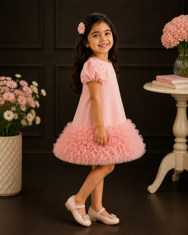 A young girl with long dark hair wearing a light pink dress with a ruffled tulle skirt and white Mary Jane shoes, standing in a decorated room with dark paneled walls and pink flowers.